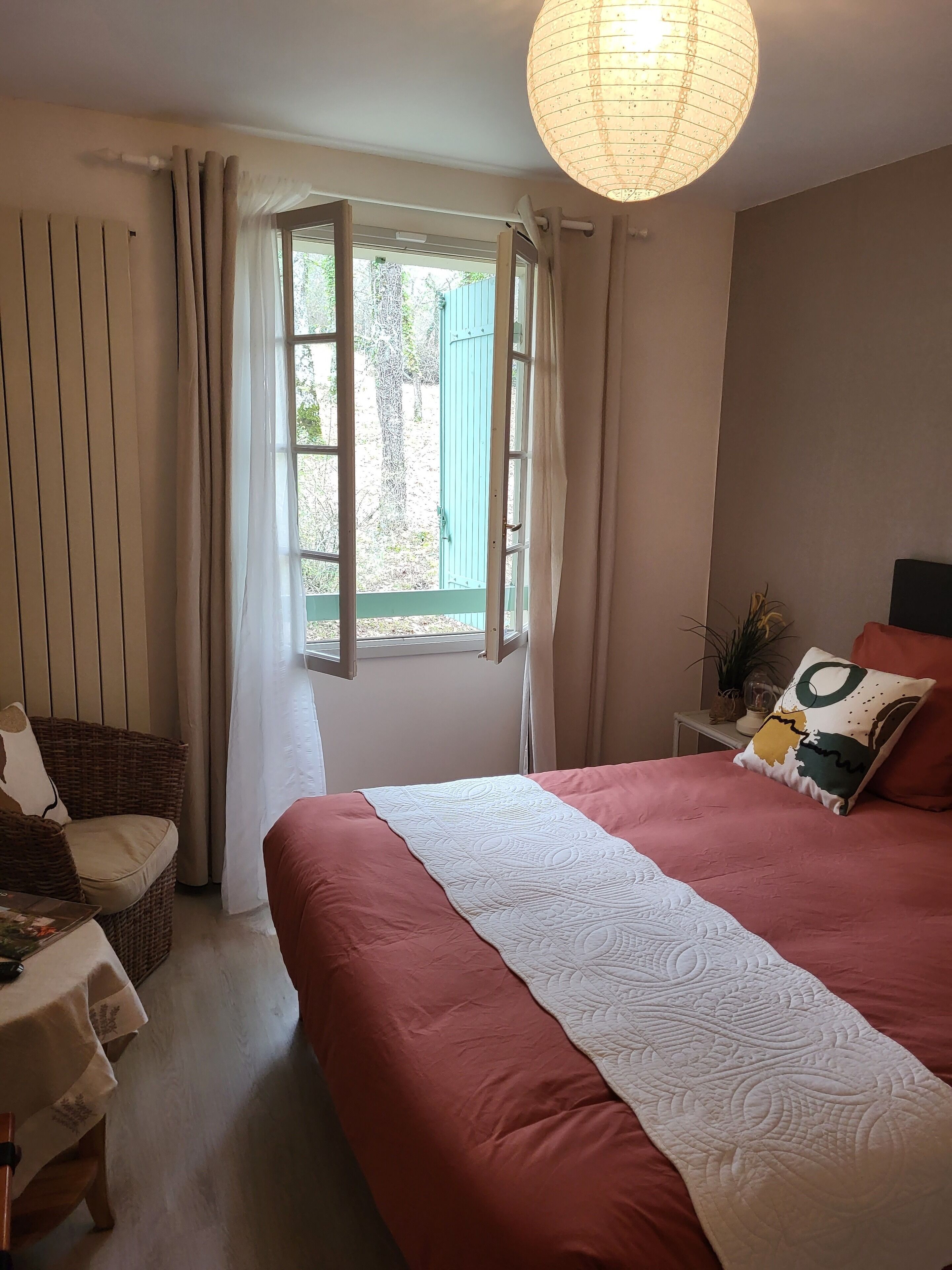 Ein Schlafzimmer mit einem großen Bett, einem Fenster mit Vorhängen, einem Nachttisch mit Lampe und einer Pendelleuchte.