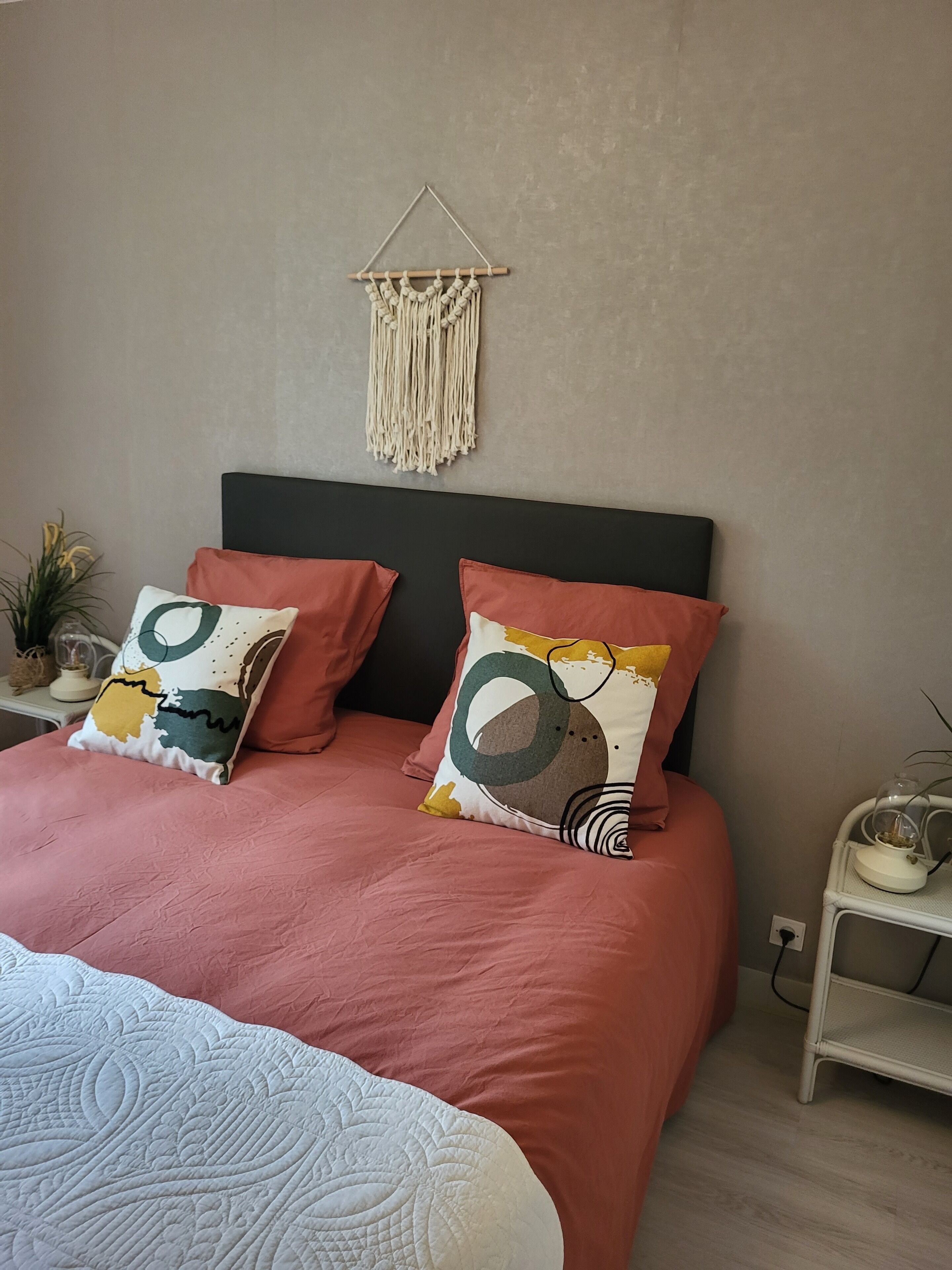 A bedroom with a bed, two pillows, a macrame wall hanging, a small plant, and a nightstand with a lamp.