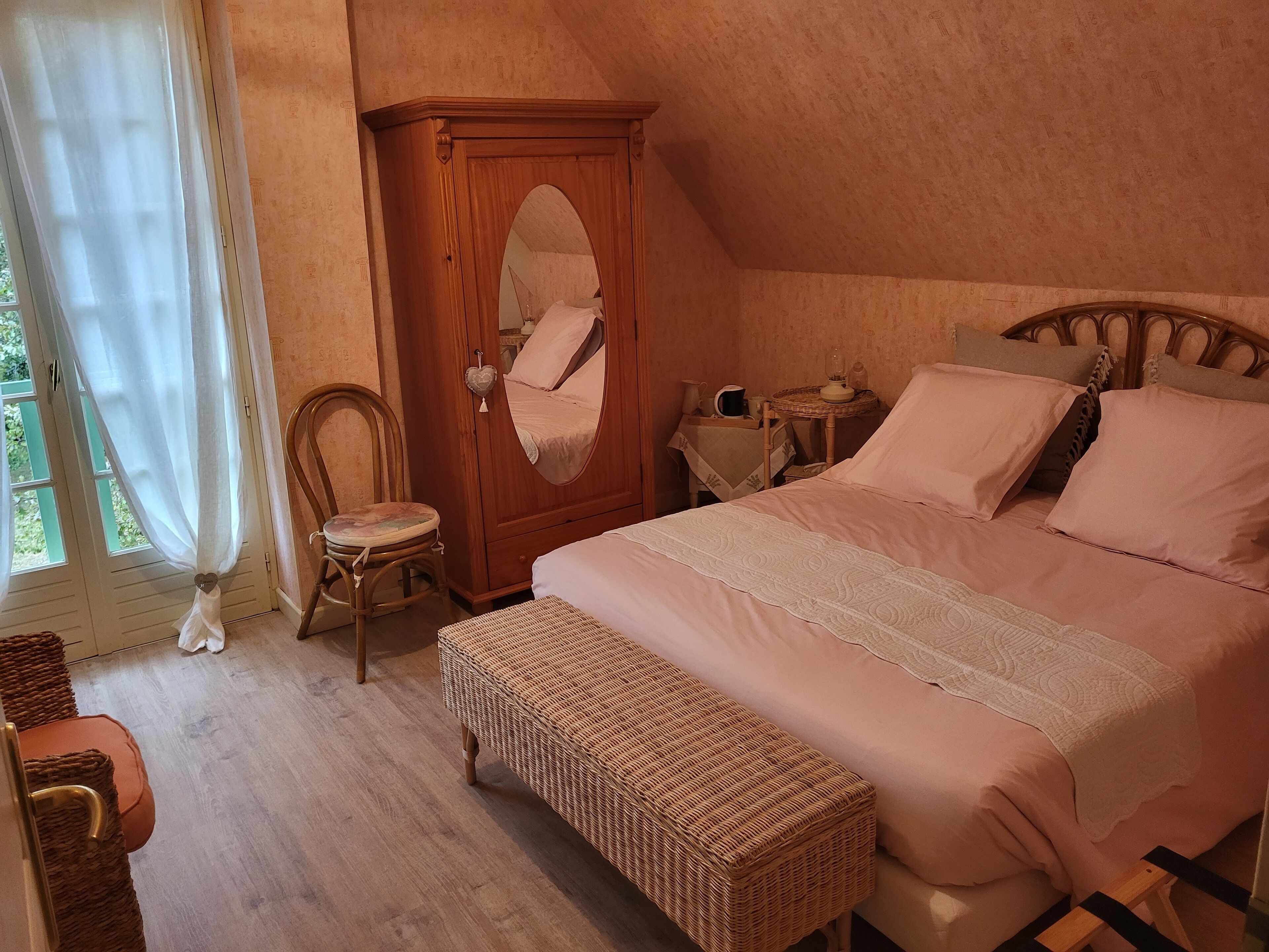 A bedroom with a bed, a wooden wardrobe, a wicker bench, a chair, and a small table with a lamp.