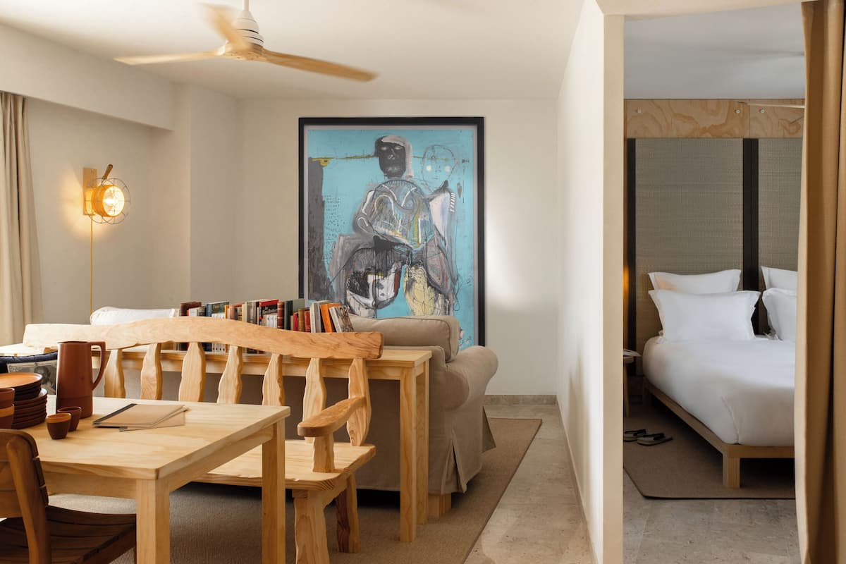 A hotel room with a bed, a desk with books, a wooden chair, and a painting on the wall.