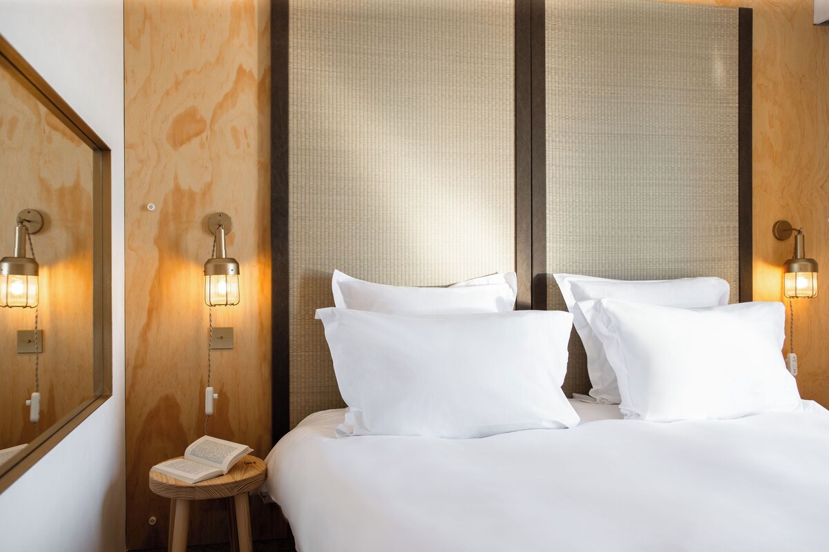 A hotel room with a neatly made bed, two bedside lamps, a small wooden stool with a book, and a wall-mounted telephone.