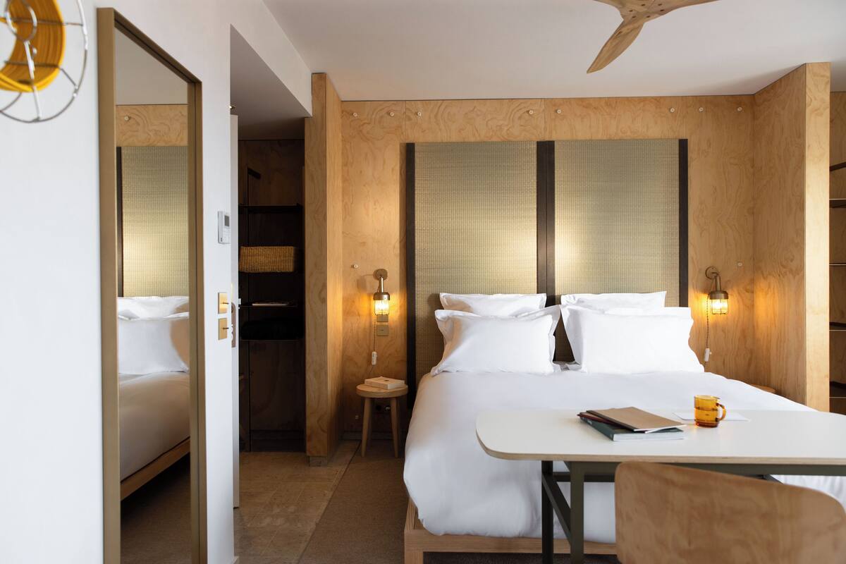 A hotel room with a large bed, a wooden headboard, two bedside lamps, a small table with a book and a mug, and a ceiling fan.