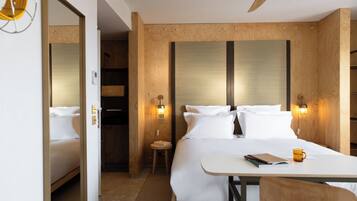 Een hotelkamer met een groot bed, een houten hoofdeinde, twee bedlampen, een klein tafeltje met een boek en een mok, en een plafondventilator.