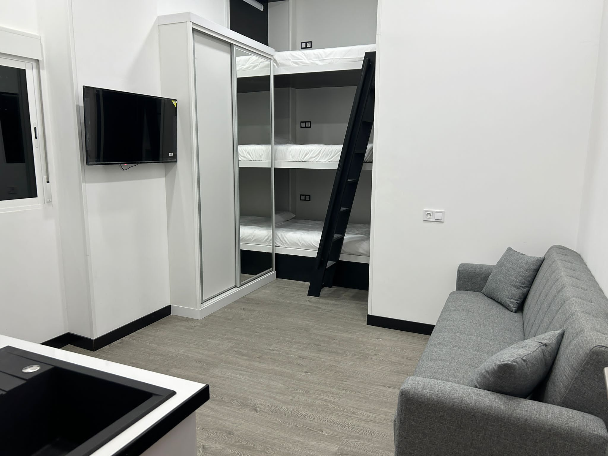 A compact room with bunk beds, a grey sofa, a TV, and a small kitchenette.