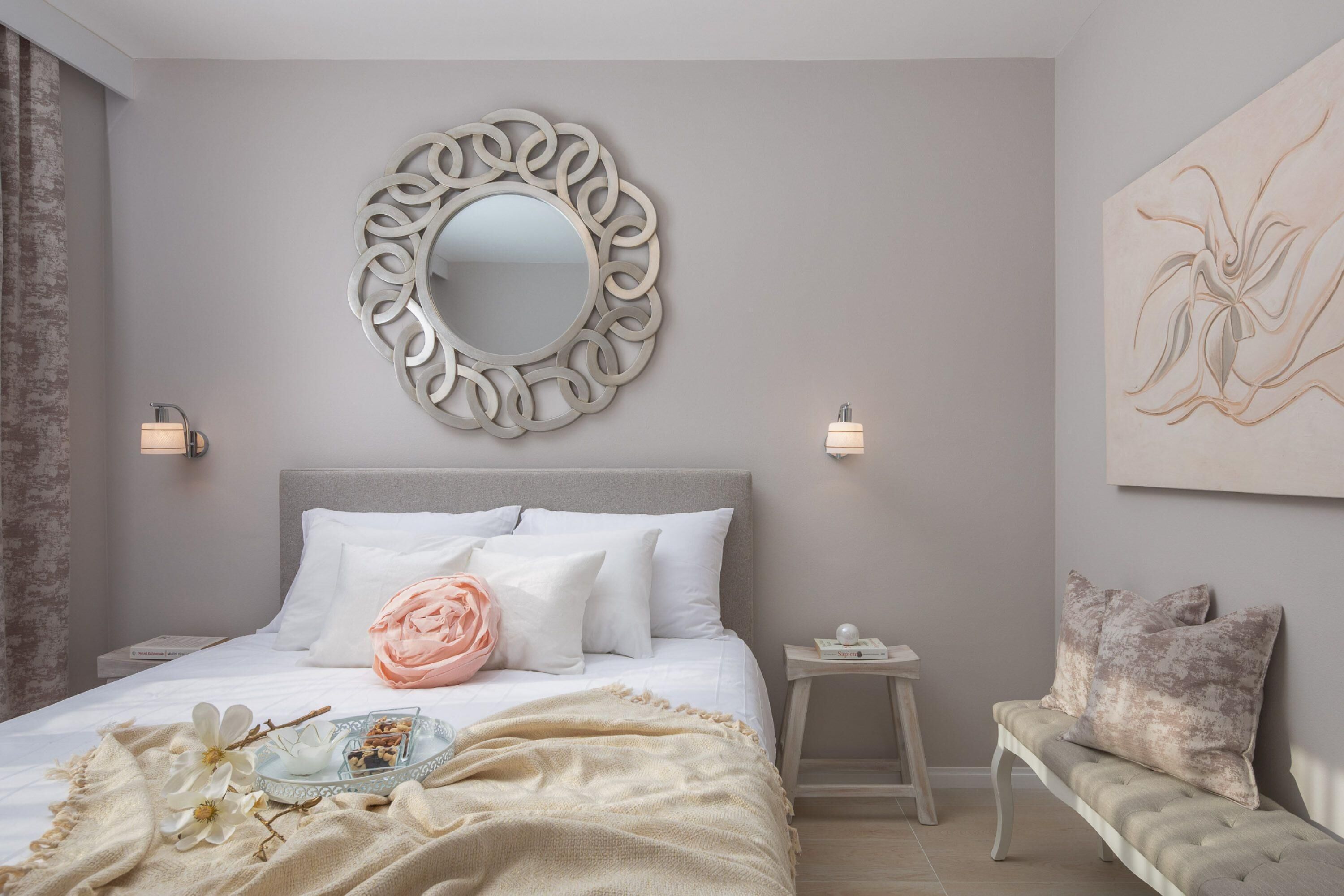 A bedroom with a large bed, a round mirror, bedside lamps, a bench, and a wall art piece.