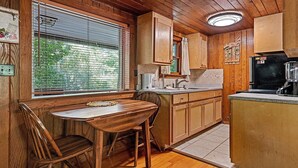 A compact kitchen with wooden cabinets, a dining table with chairs, a window with blinds, and a refrigerator. - Sunflower Shack: 1  BR, 1  BA Cabin / Bungalow in Maggie Valley, Sleeps 2 (Maggie Valley)