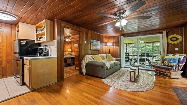 A cozy cabin interior with wooden walls, a kitchen area, a living room with a sofa and a coffee table, and a large window with a view of greenery. - Sunflower Shack: 1  BR, 1  BA Cabin / Bungalow in Maggie Valley, Sleeps 2 (Maggie Valley)