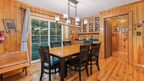 A wooden interior with a dining table and chairs, a kitchen area with cabinets, and a large window with curtains. - Huckleberry Hideaway (Maggie Valley)