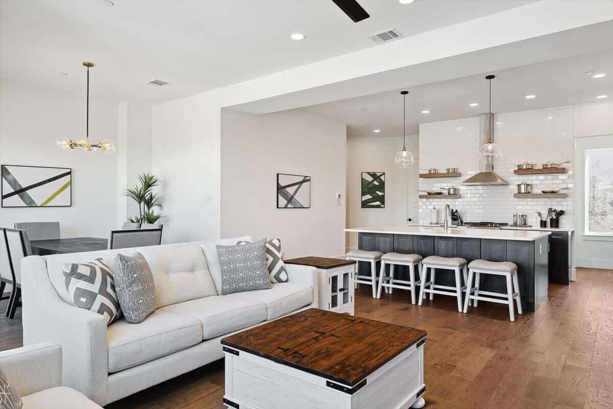 Una sala moderna con sofá, mesa de centro y taburetes de bar. Se ve una cocina con isla central e iluminación colgante al fondo.