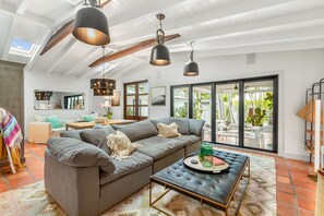 A spacious living room with a gray sectional sofa, a leather ottoman, and a dining area with a wooden table. Large windows provide ample natural light and a view of the outdoors. - SUN DAY, FUN DAY ~ Renovated 2/2 Home 2 Blocks from Beach in Casa Marina! (Key West)