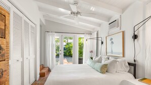 Een slaapkamer met een groot bed, witte muren, een plafondventilator en uitzicht op een tuin door een schuifdeur.