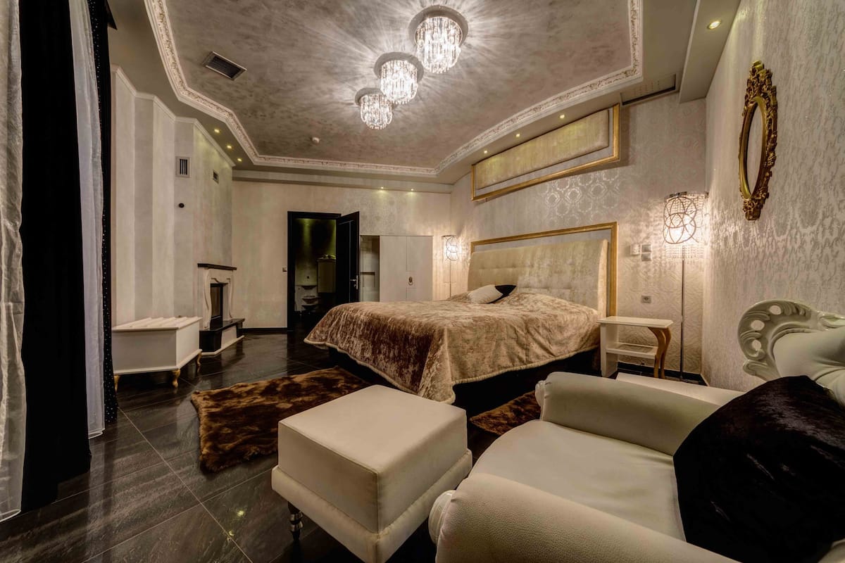 Une chambre à coucher luxueuse avec un grand lit, un coin salon avec un canapé et un pouf, une coiffeuse avec un miroir et un lustre.