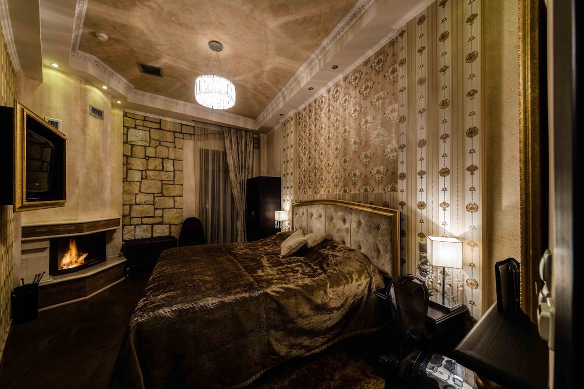 Une chambre d’hôtel avec une cheminée en pierre, un grand lit recouvert de velours, un lustre et un papier peint décoratif.