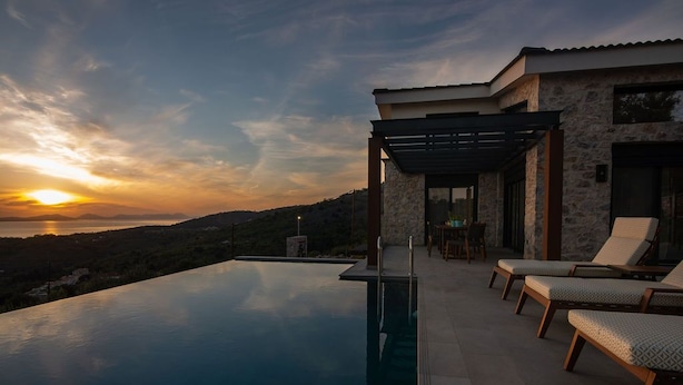 An infinity pool with a view of the sunset, outdoor seating, and a stone building.