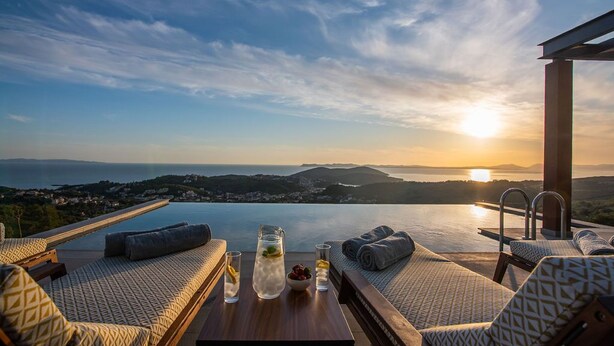 A sunlit terrace with lounge chairs, a table with drinks, and a scenic view of the sea and hills at sunset. - GV Sivota Luxury Villa - SL2021 (Igoumenitsa)