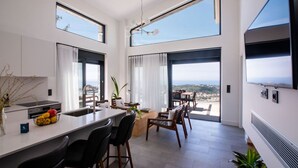 A modern kitchen with a central island, dining area with chairs, large windows, and a view of the landscape. - GV Sivota Luxury Villa - SL2021 (Igoumenitsa)