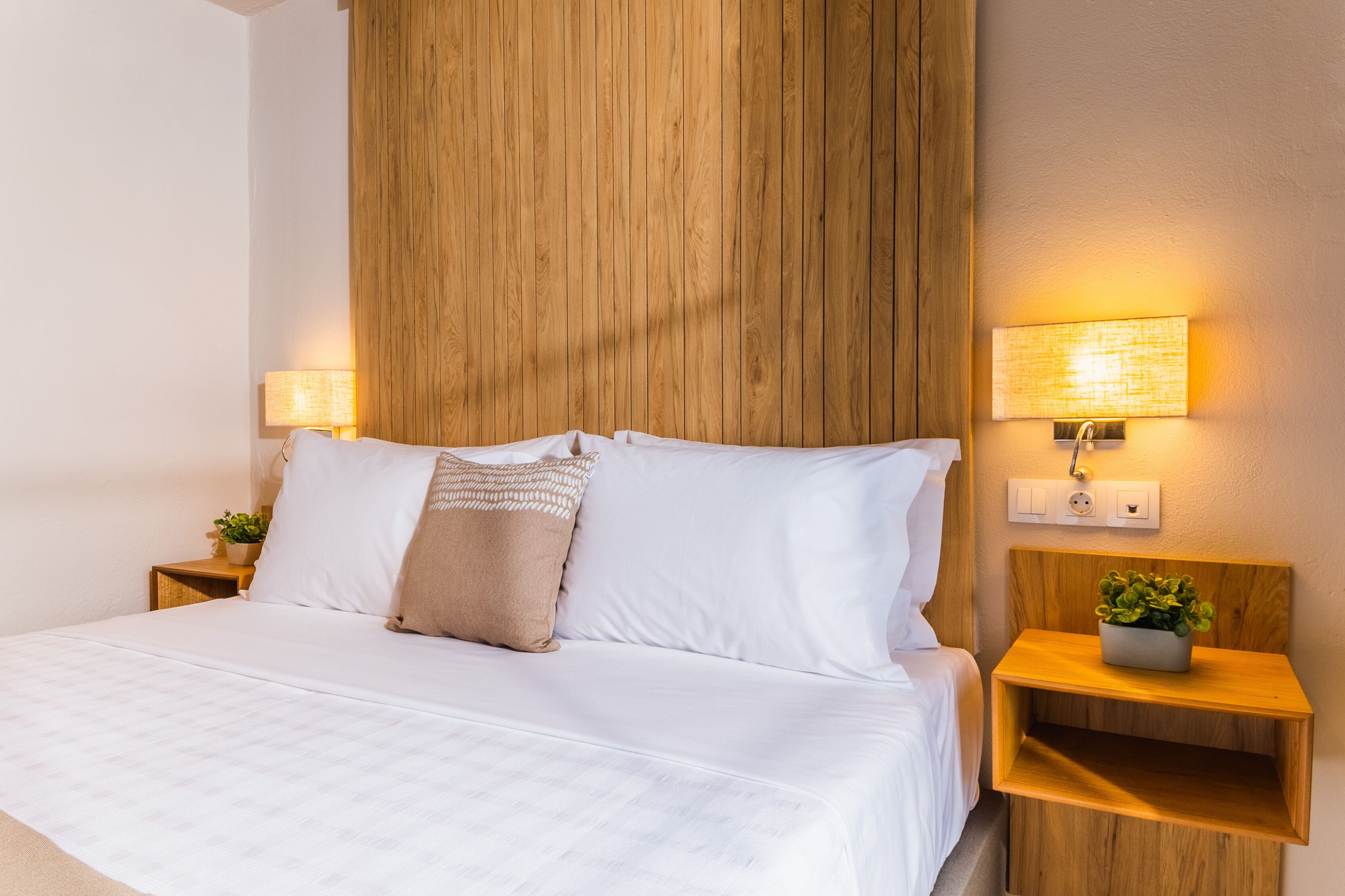 Een netjes opgemaakt bed met witte lakens en een beige sierkussen, aan weerszijden omringd door houten nachtkastjes met lampen en kleine potplanten.