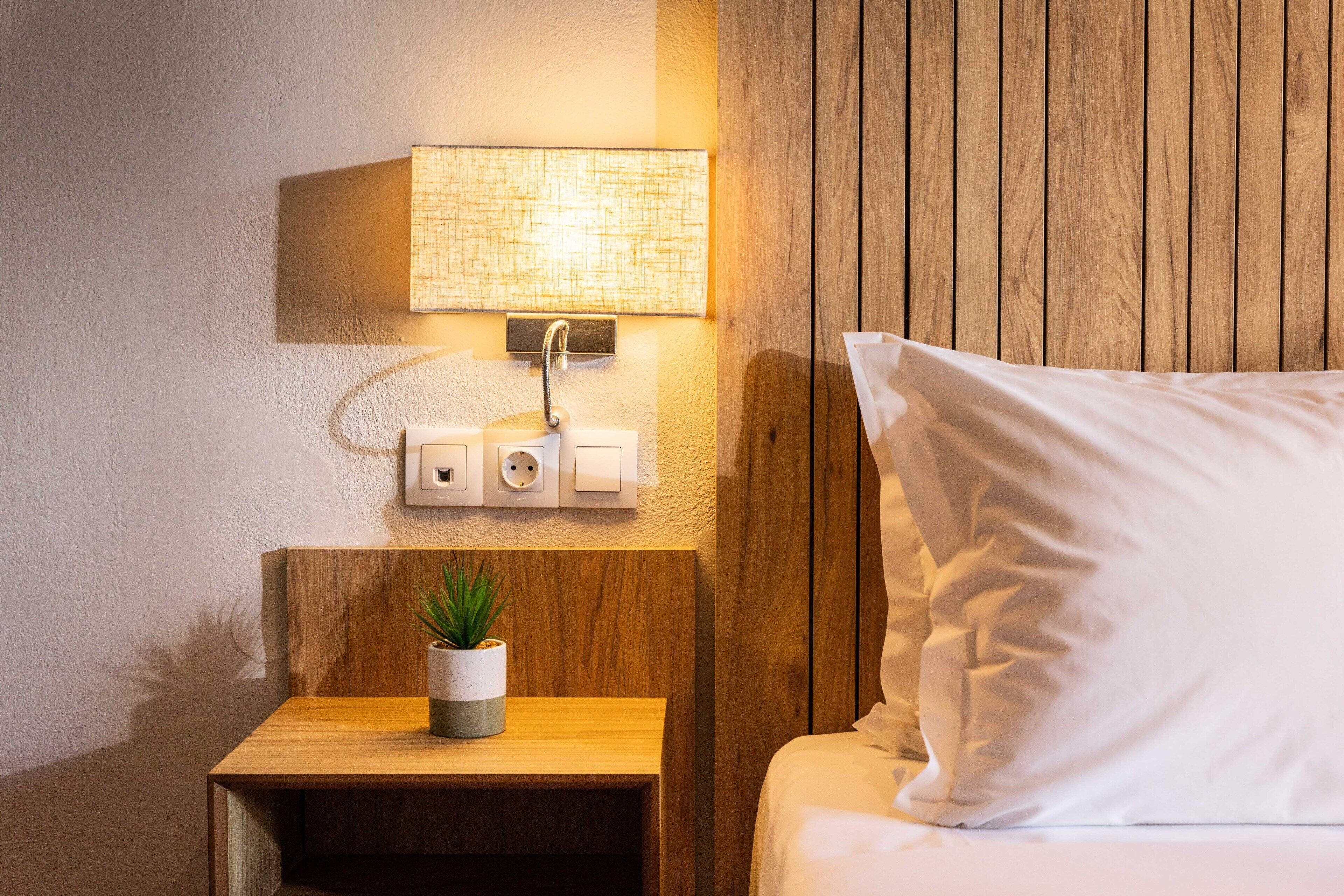 A neatly made bed with white linens, a bedside lamp, and a small wooden nightstand with a potted plant.