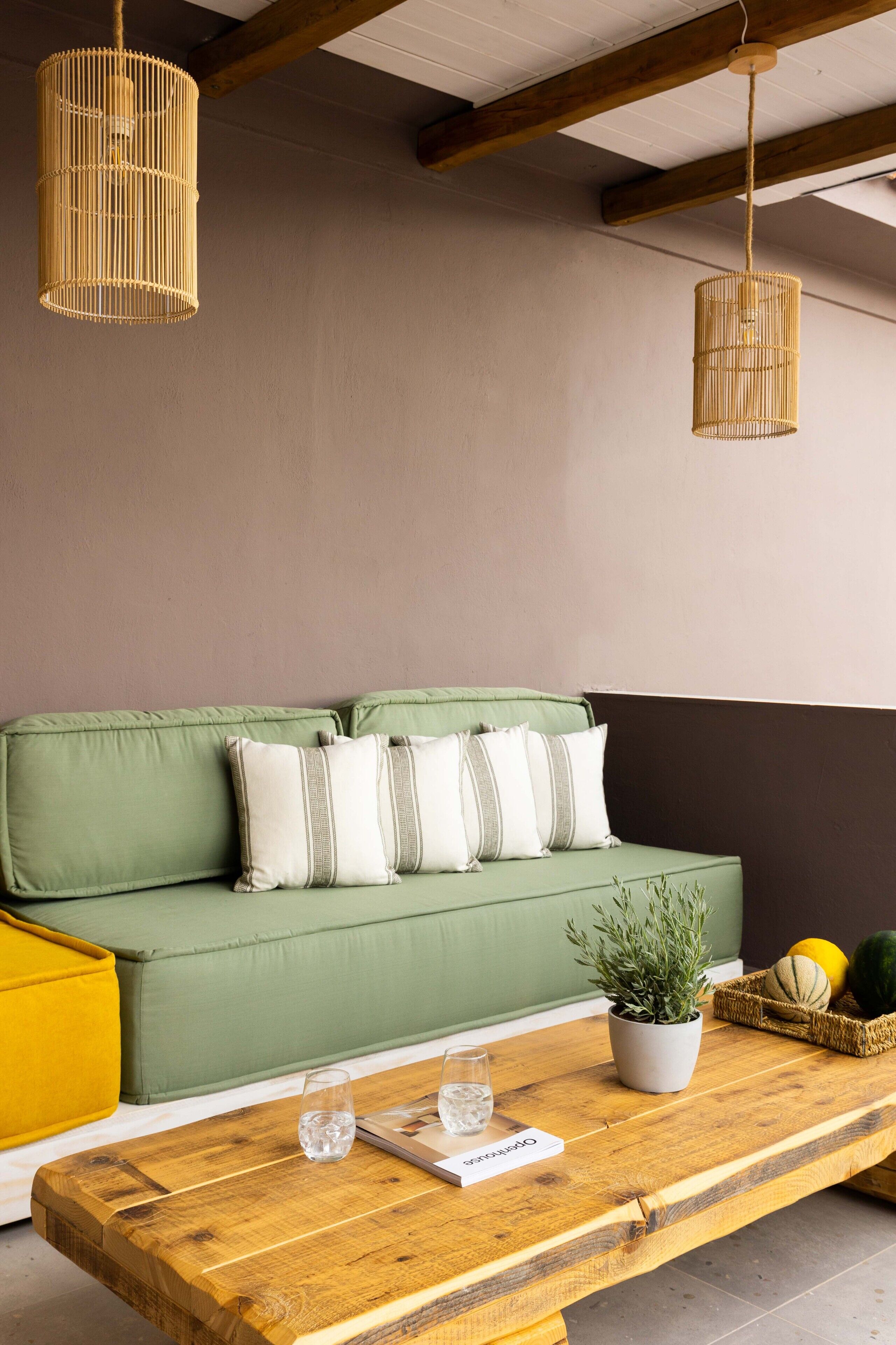 A green sofa with striped pillows, a wooden coffee table with a plant and a basket of fruit, and two hanging wicker lamps.