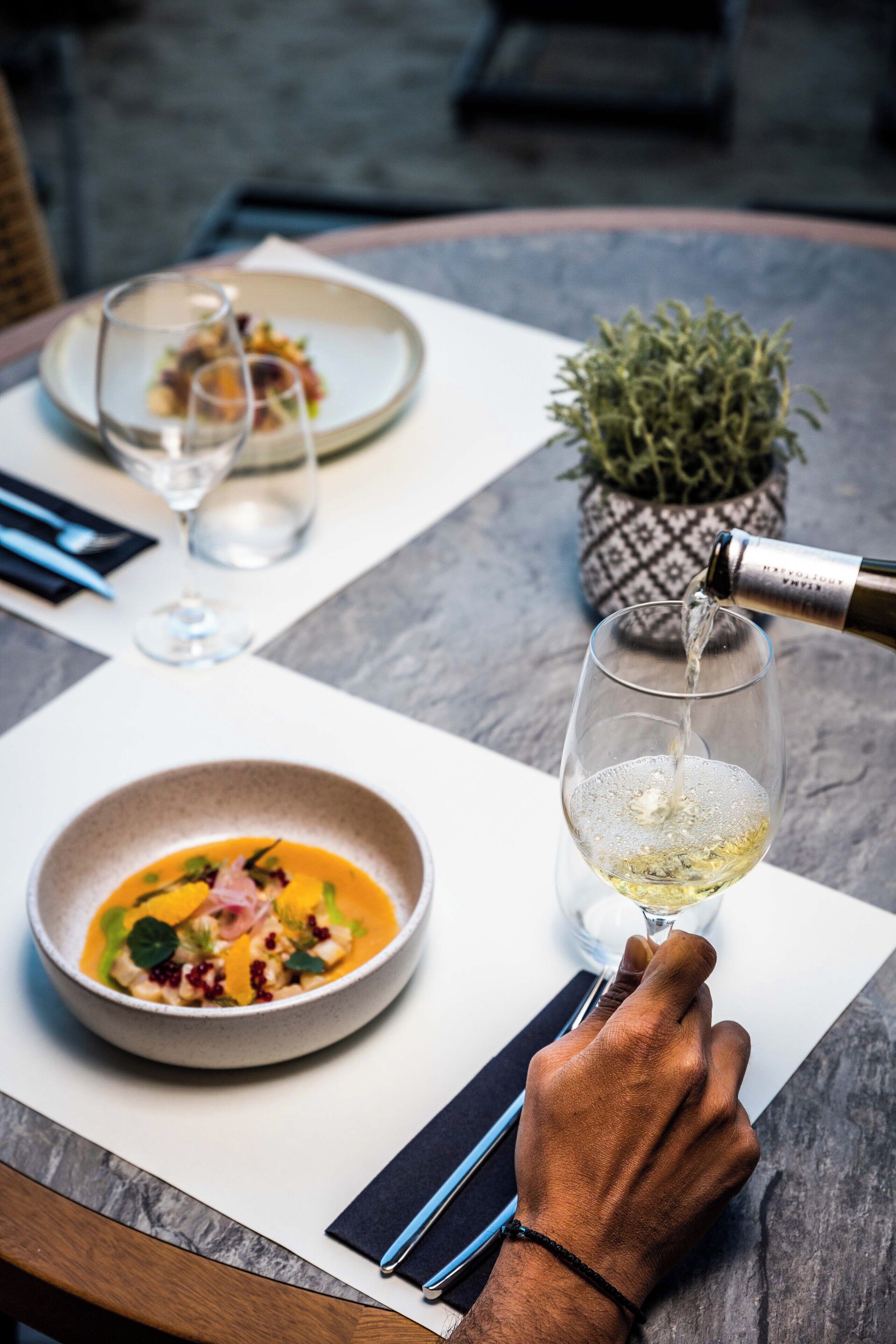 A dining table with a bowl of soup and a glass of white wine being poured.