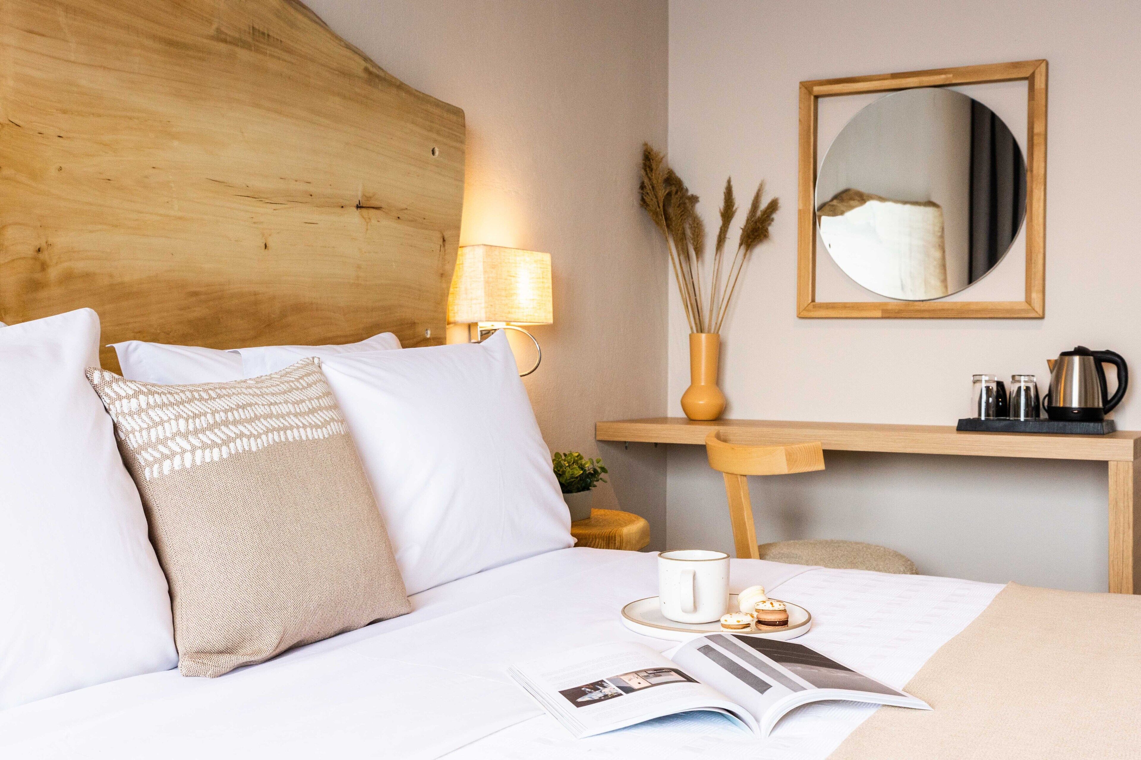 A neatly made bed with white and beige pillows, a bedside lamp, a small table with a cup and a book, and a mirror on the wall.