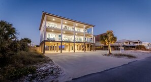 A multi-story hotel with a covered outdoor area, a sandy beach in the foreground, and palm trees. - 🌴🌞Private Pool + Stunning Gulf Views | Perfect for Groups | Emerald Oasis🌴🌞 (Pensacola Beach)