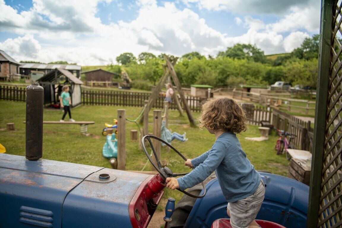 Niño conduciendo un tractor en un área de juegos rural con estructuras de madera y una valla.