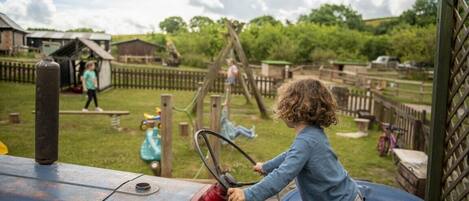 Niño conduciendo un tractor en un área de juegos rural con estructuras de madera y una valla.
