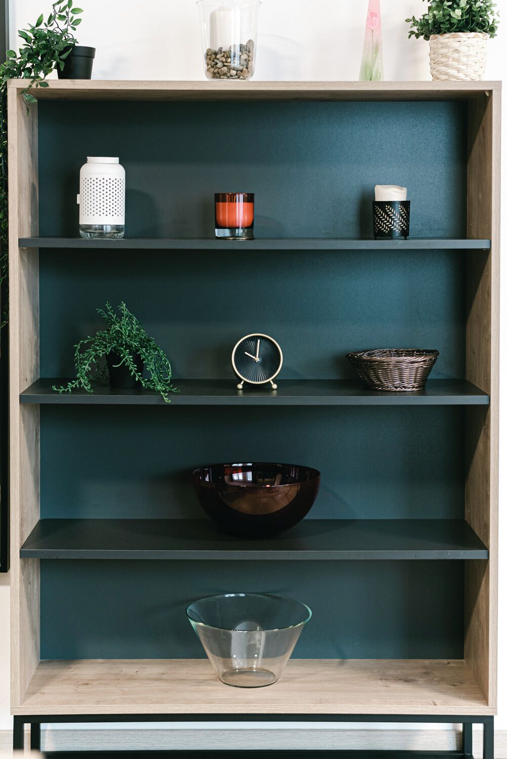 A wooden shelf with various items including a white perforated container, a candle, a small clock, a woven basket, and a clear glass bowl.