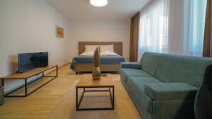Een moderne hotelkamer met een bed, een groene bank, een televisie op een houten standaard en een vaas op een bijzettafeltje.