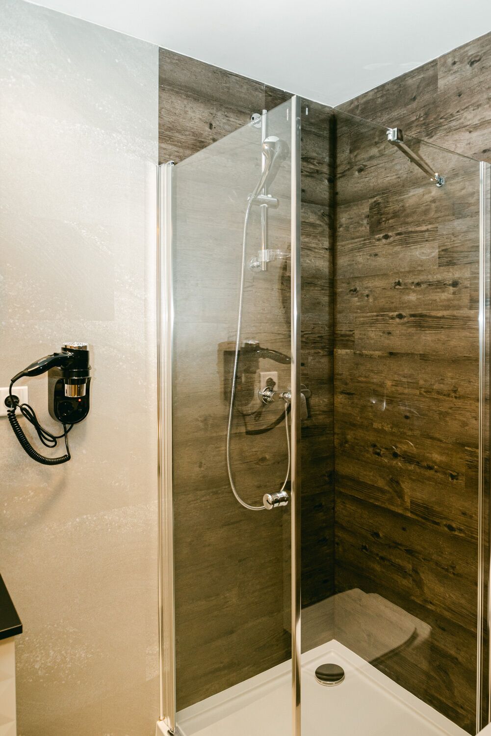 A glass-enclosed shower with a wooden wall panel and a black hairdryer on the wall.