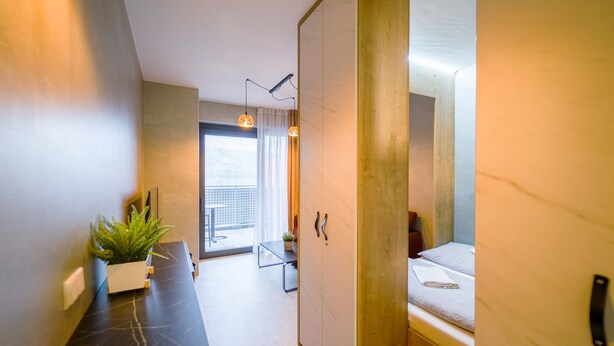 A modern hotel room with a bed, a desk with a potted plant, a wardrobe, and a view of the outdoors. - Svarog Unique Apartment by CHORS (Bratislava)