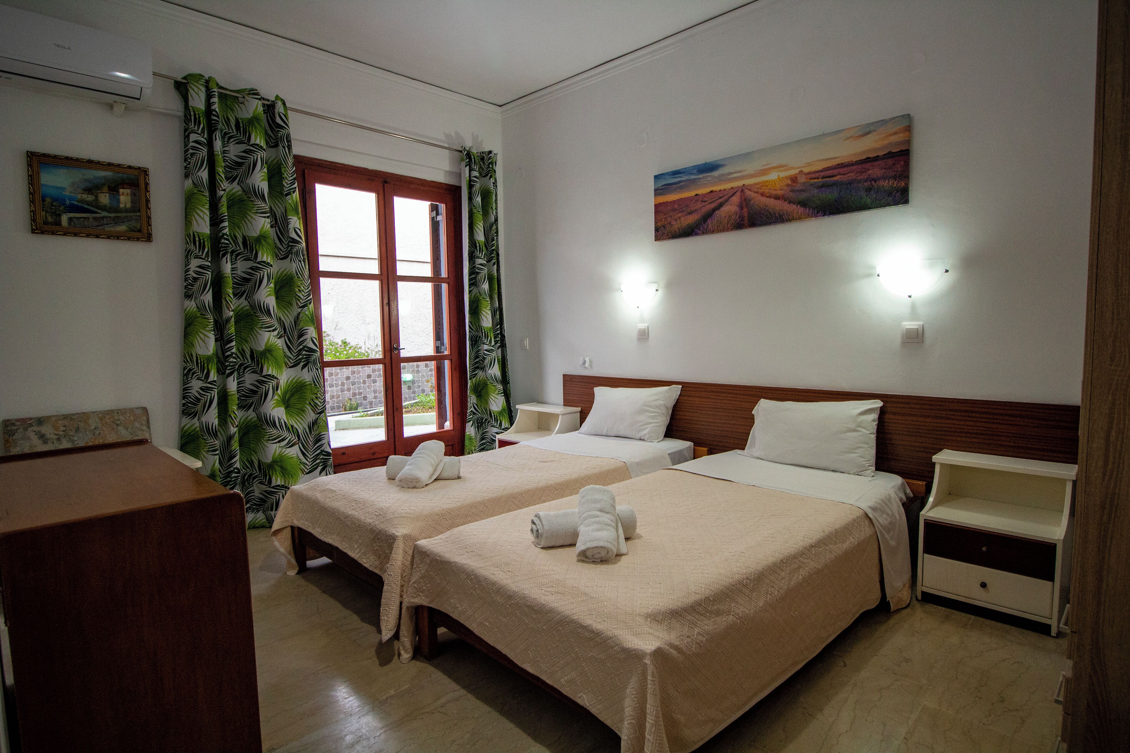 A hotel room with two beds, a wooden desk, a nightstand, a painting, and a window with curtains.
