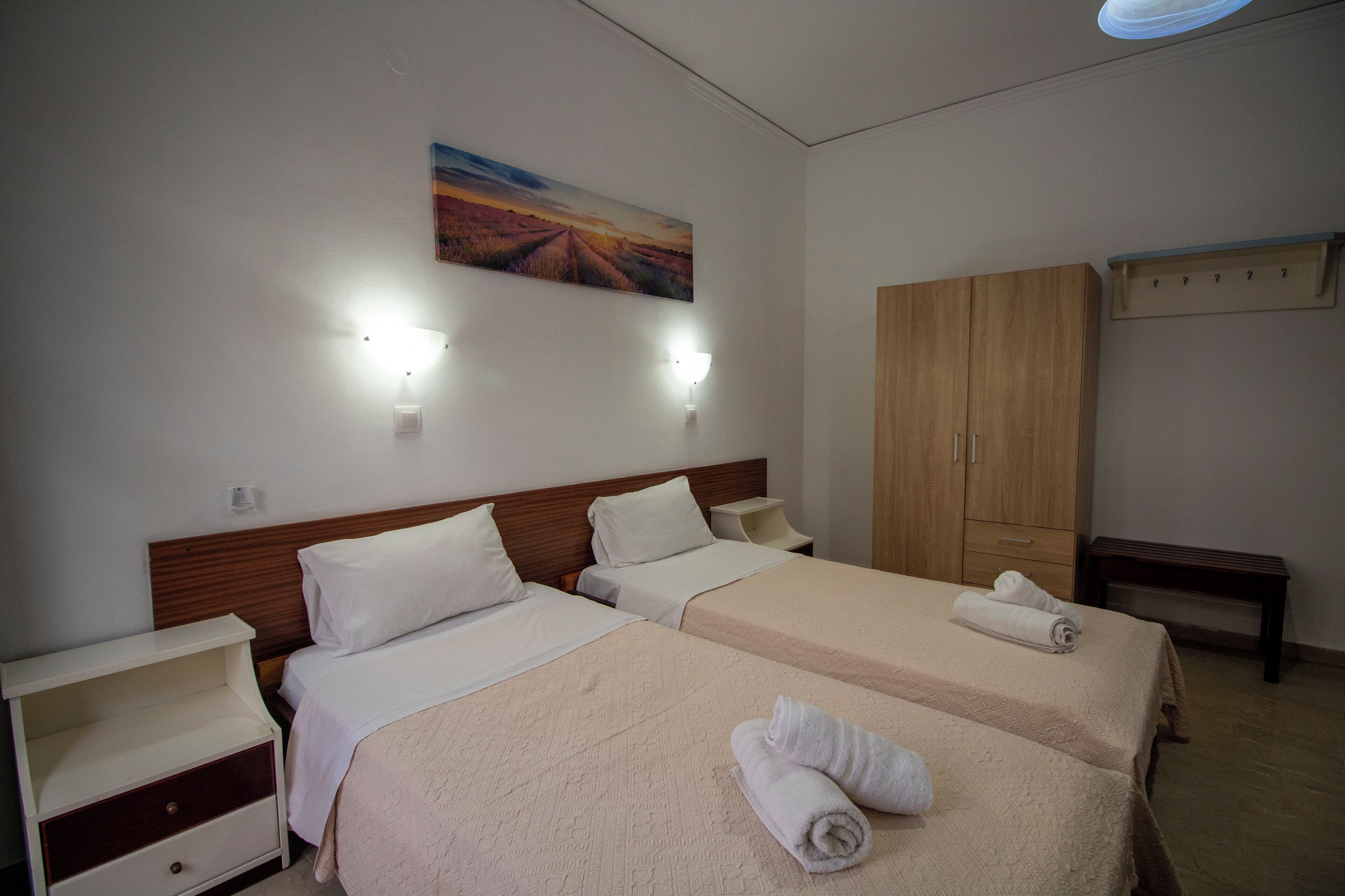 A hotel room with two beds, a wooden wardrobe, a bench, and a wall-mounted light fixture.