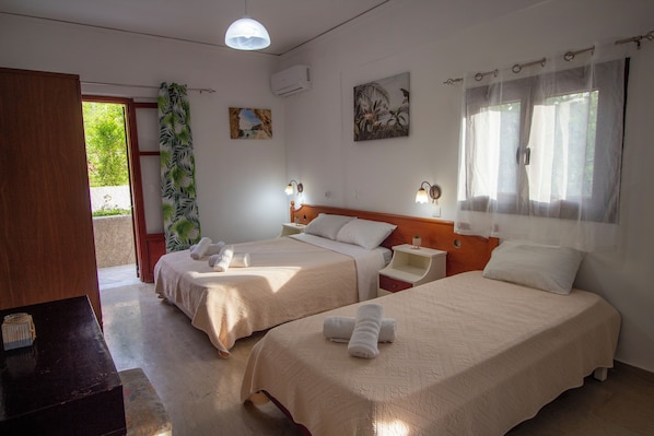 A hotel room with two beds, a wooden headboard, a nightstand with a lamp, a small table, and a door leading outside.
