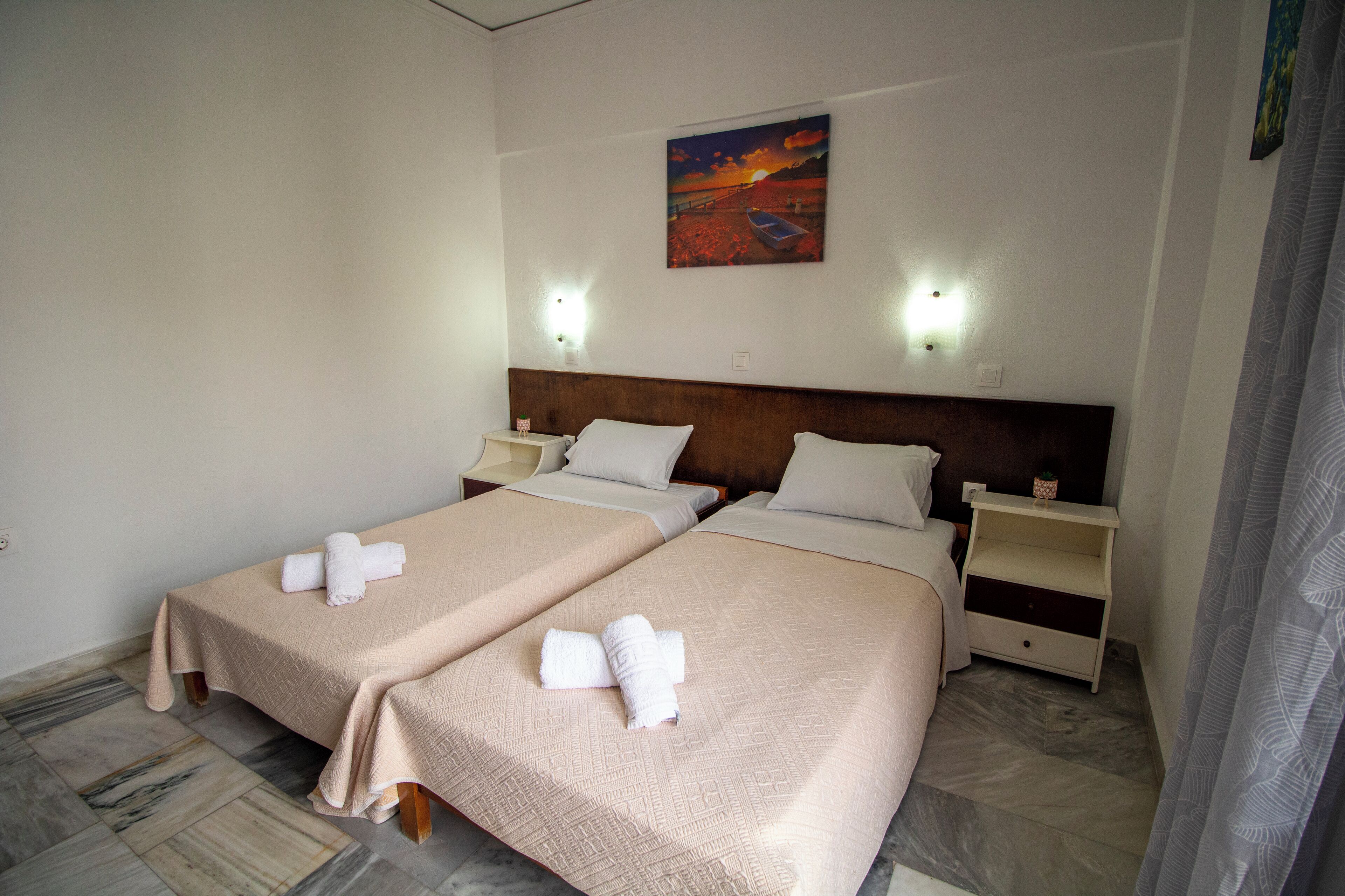 A hotel room with two beds, each with a beige bedspread and white pillows. There are two bedside tables with lamps, a painting on the wall, and a patterned floor.