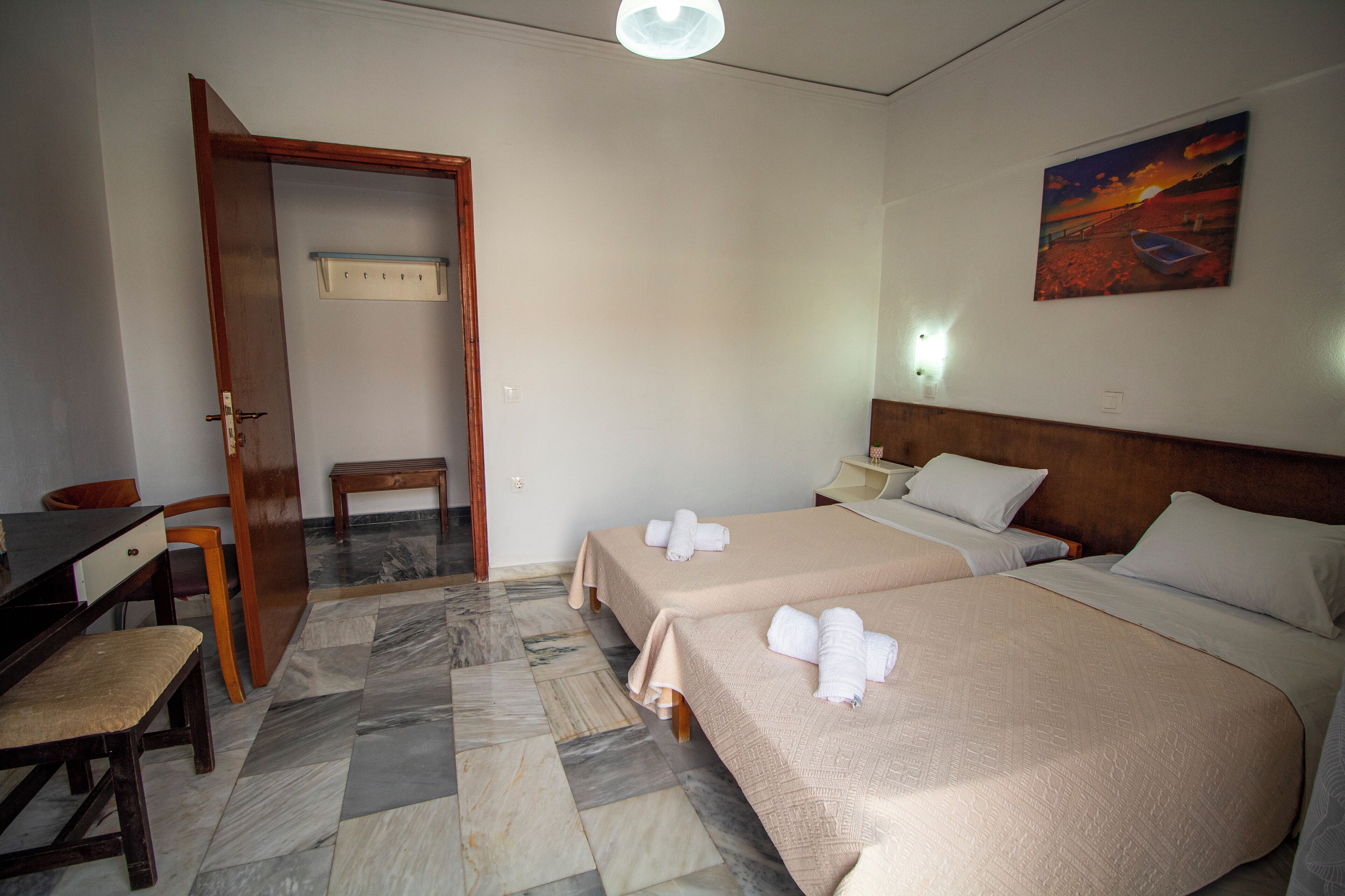 A hotel room with two beds, a desk, a chair, a wooden door, a bench, and a painting on the wall.