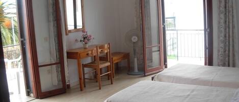A hotel room with two beds, a desk with a chair, a fan, and a window with a view of the outside.