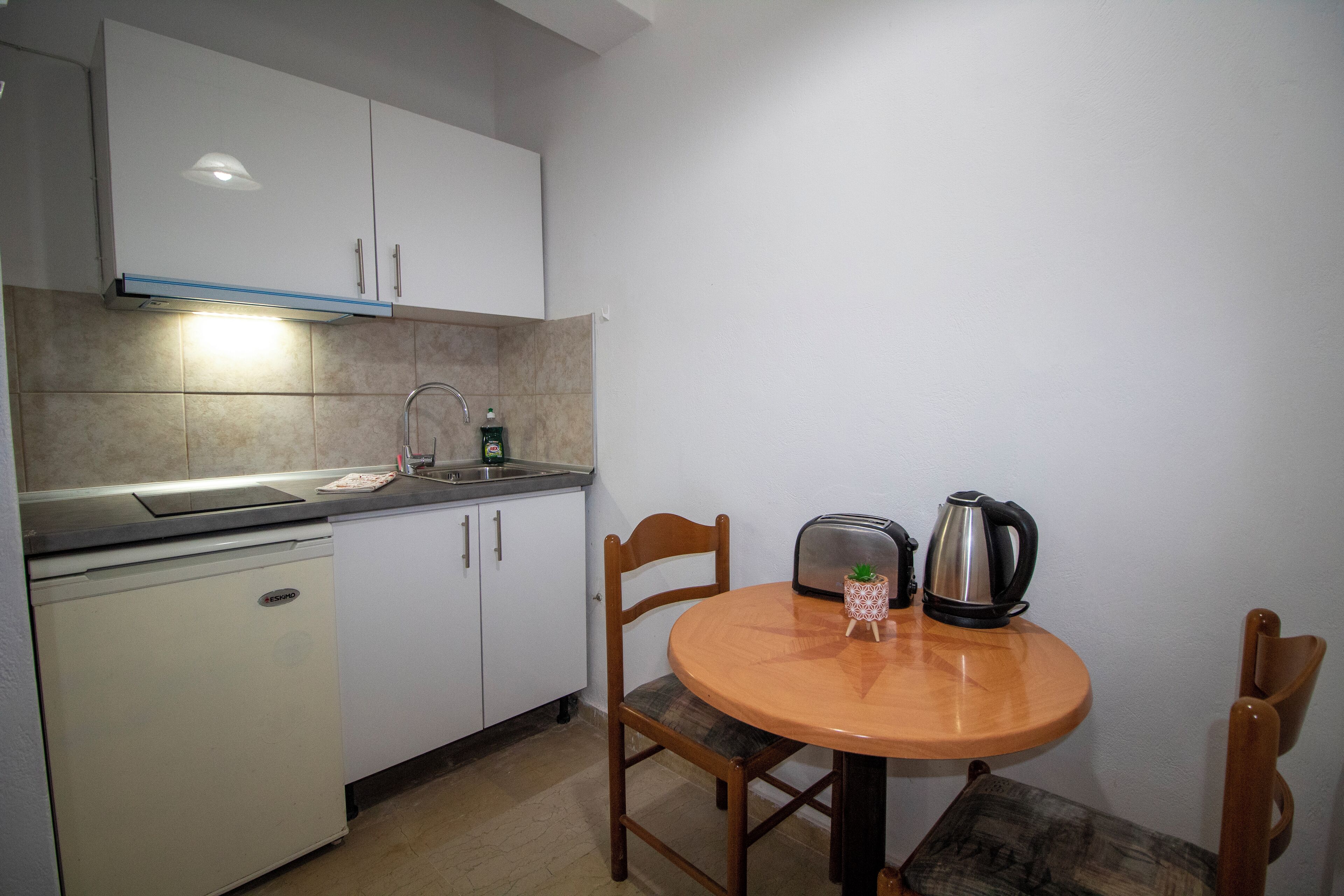A compact kitchen with white cabinets, a built-in dishwasher, a round wooden table with two chairs, a toaster, and a kettle.