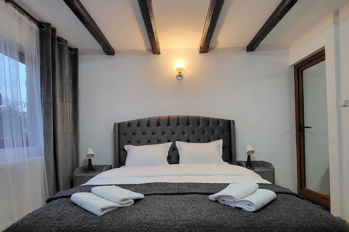 A hotel room with a large bed, two bedside tables with lamps, a ceiling light, and a view of the outside through a window with curtains.