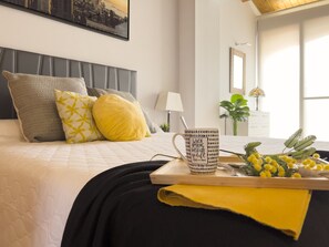 A neatly made bed with pillows, a tray with a mug and flowers, and a cityscape painting on the wall.