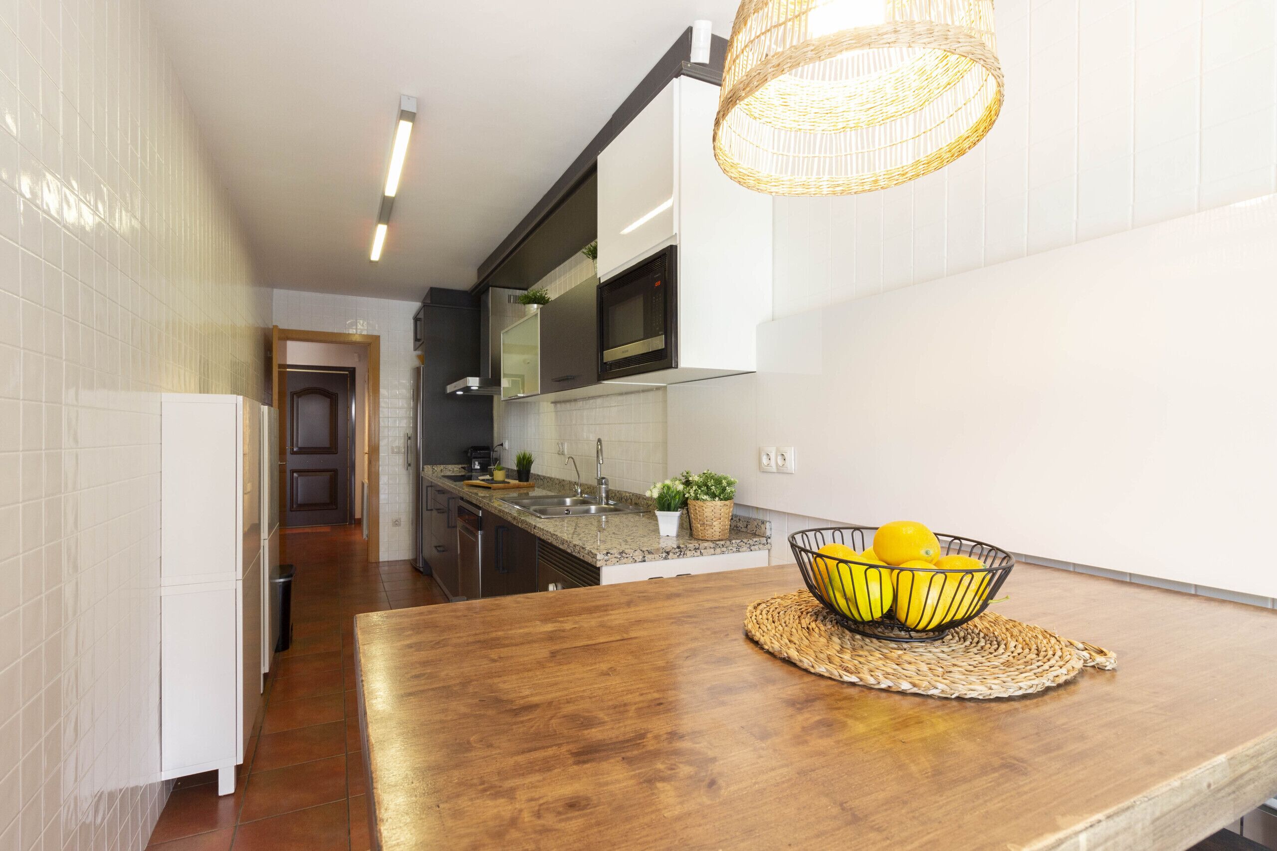 A modern kitchen with white tiles, a wooden countertop, a woven basket with lemons, and a hanging light fixture.