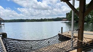 Una hamaca en un muelle de madera con vista a un lago tranquilo, con un barco amarrado y una orilla lejana.
