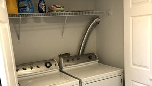 A laundry room with two white washers and dryers, a metal shelf above holding cleaning supplies, and a vent pipe. - Bright open studio apartment with king bed and queen sofabed. Walk to local shops and restaurants. (Hickory)
