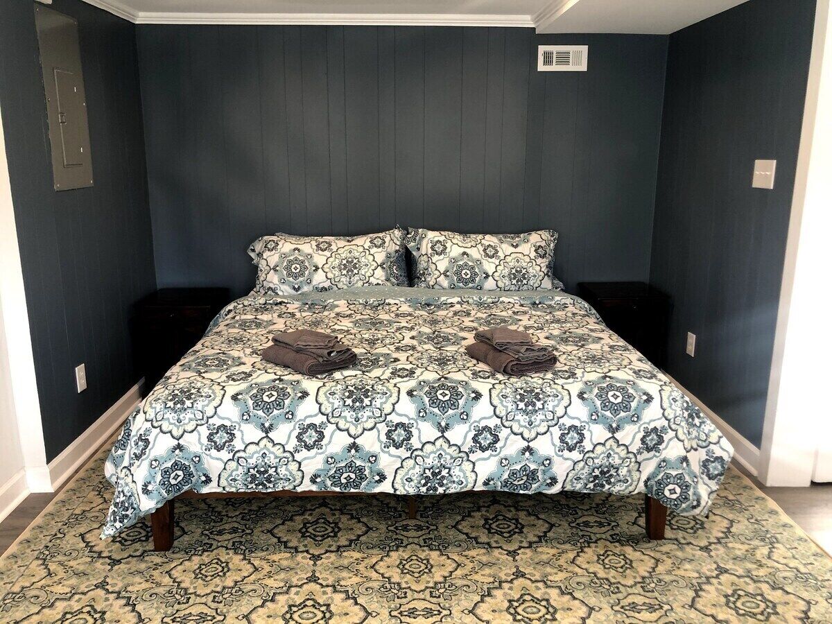 A bed with a patterned bedspread and two pillows, a dark wall, a small window, and a door.