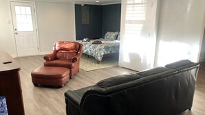 A modern living room with a black leather sofa, a brown leather chair, and a small ottoman. There is a bed with a patterned bedspread in the background. - Bright open studio apartment with king bed and queen sofabed. Walk to local shops and restaurants. (Hickory)