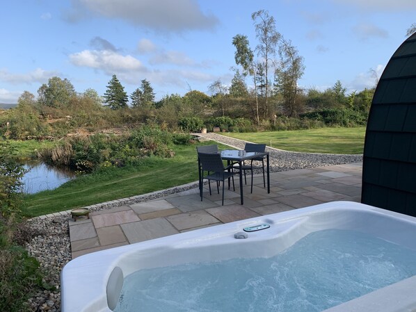 An outdoor hot tub area with a table and chairs, surrounded by a landscaped garden and a pond. - Red Squirrel Pod with Hot Tub - perfect for relaxing in countryside near Oban (Oban)