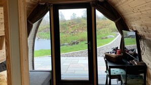 A cozy cabin interior with a stone fireplace, a comfortable seating area, and a scenic view of a lake and garden through a large glass door. - Red Squirrel Pod with Hot Tub - perfect for relaxing in countryside near Oban (Oban)