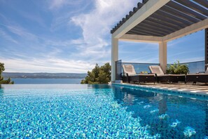 Una piscina infinita con vistas despejadas al mar, un área de estar techada con sillones reclinables y un diseño arquitectónico moderno.