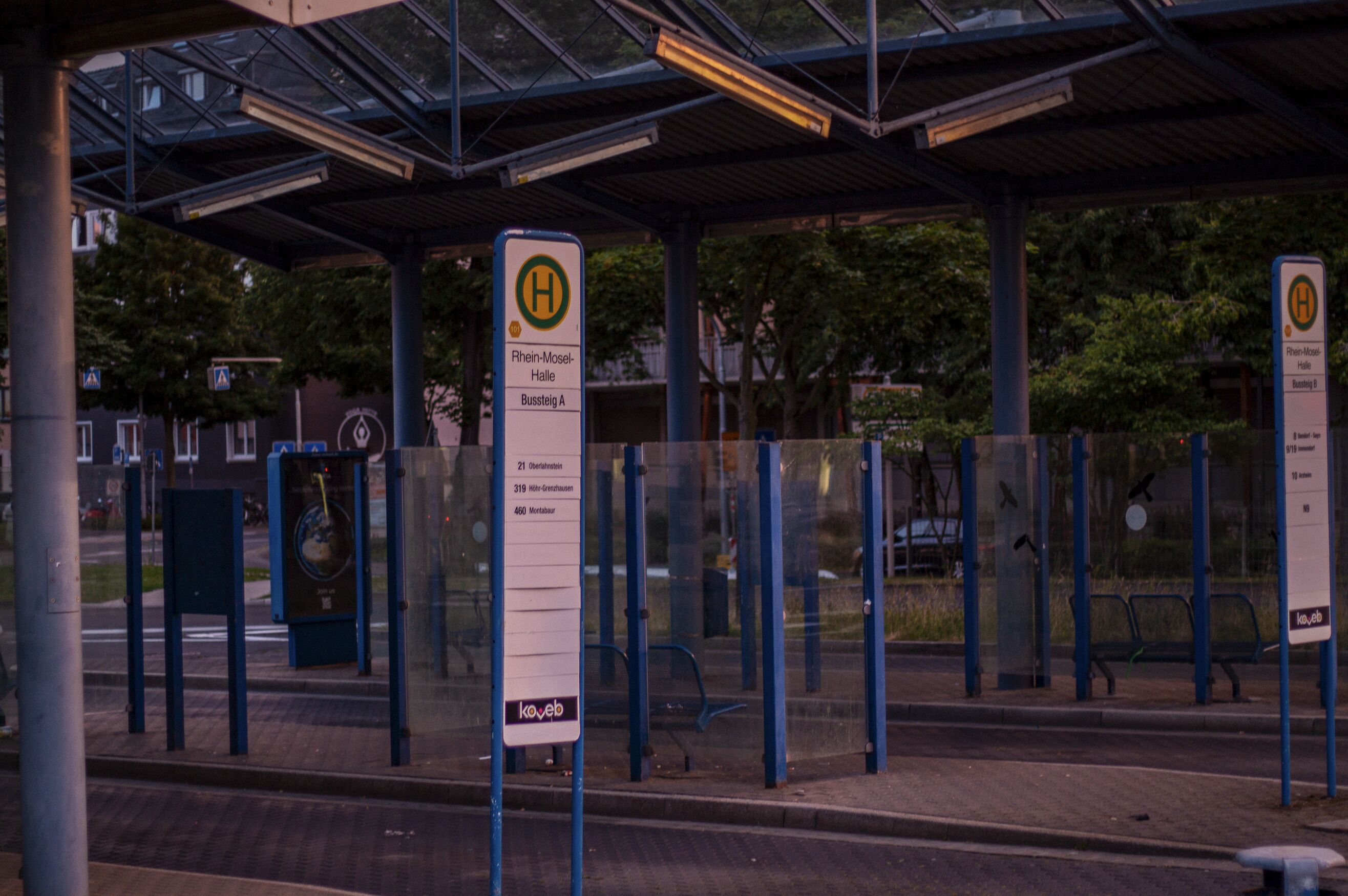 Eine Bushaltestelle mit Unterstand, ein Schild mit der Aufschrift 'Rhein-Mosel-Halle Bushaltestelle A' und ein 'koyeb'-Logo.